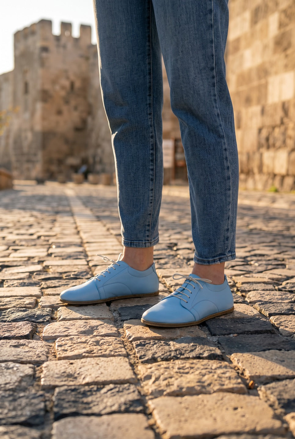 Person in light blue jeans wearing baby blue leather barefoot sneakers by Bespoky on a cobblestone path beside old stone castle walls