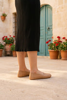 Woman in a black knit midi skirt wearing natural leather barefoot ballet flats by Bespoky in a stone courtyard with red geraniums and a turquoise door