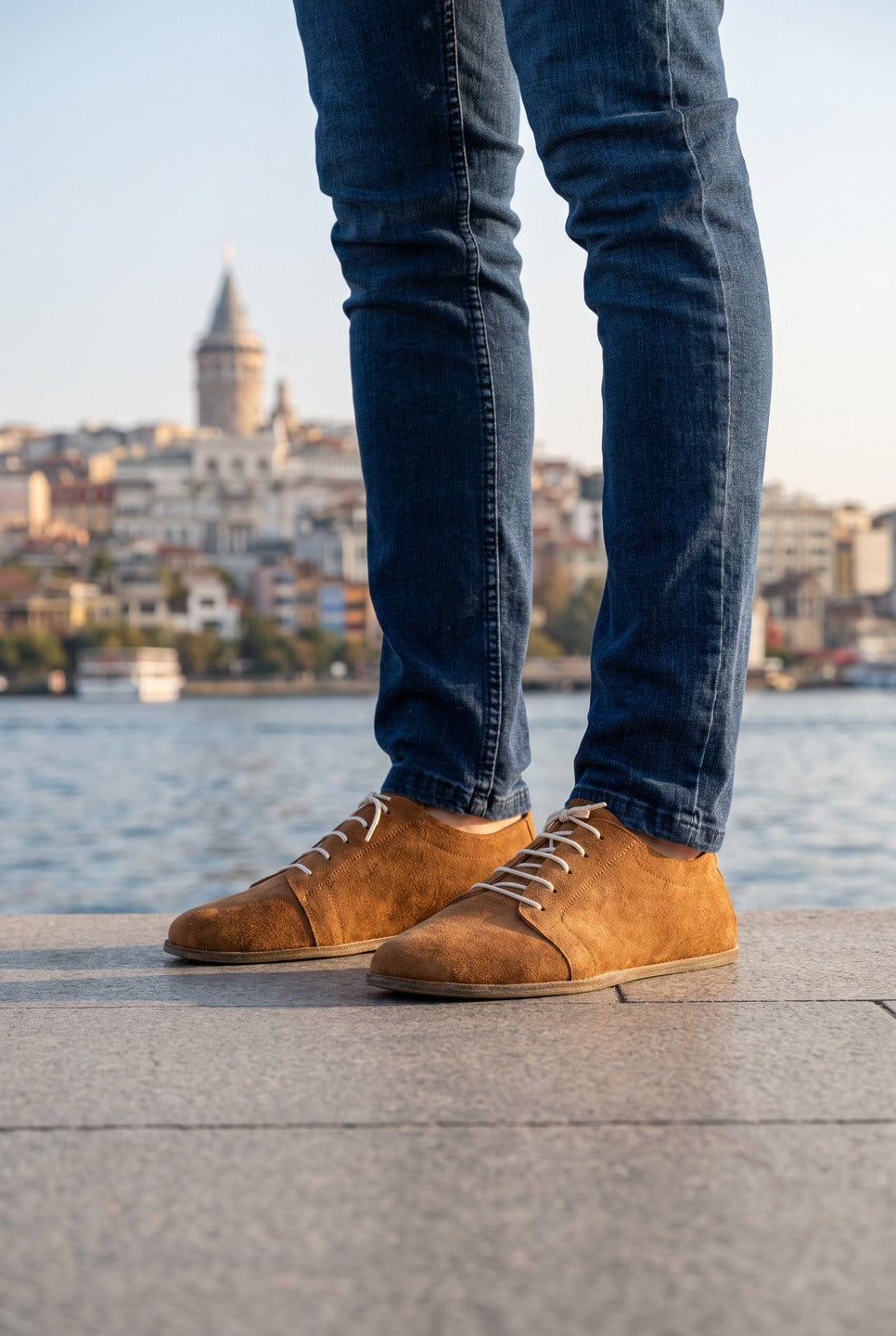 Man in dark slim jeans wearing tan suede barefoot sneakers by Bespoky on a stone waterfront with the Galata Tower in the background