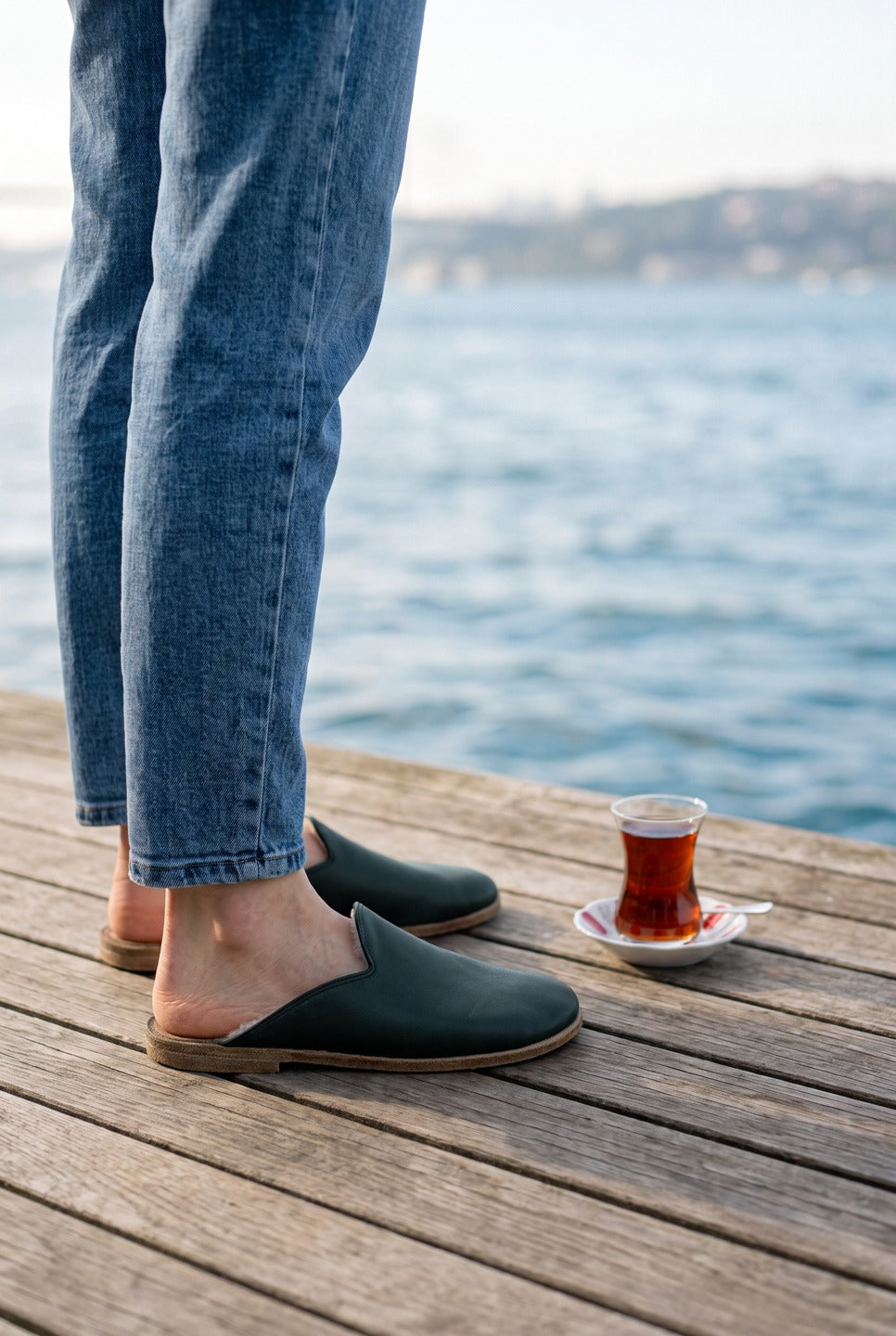 Person in light jeans wearing dark green leather shearling slippers by Bespoky on a wooden terrace by the sea next to a glass of Turkish tea