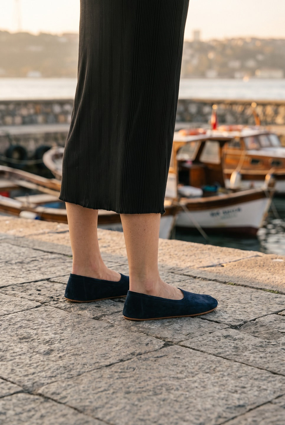 Woman in a black knit midi skirt wearing navy suede barefoot ballet flats by Bespoky on a stone seaside promenade with fishing boats in Istanbul