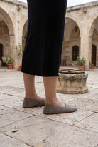 Woman in a black knit midi skirt wearing dark gray suede barefoot ballet flats by Bespoky in a limestone courtyard with arches and an old stone well