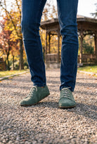 Man in dark jeans wearing green suede barefoot sneakers by Bespoky on a gravel path in an autumn park with a wooden pavilion