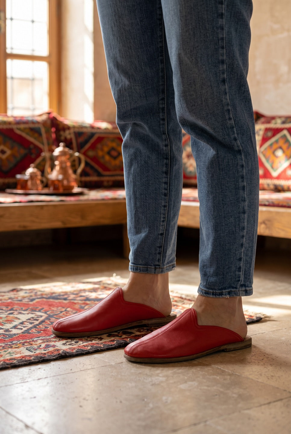 Person in light jeans wearing red leather barefoot slippers by Bespoky on a kilim rug in a traditional stone home with kilim cushions and a copper tea set