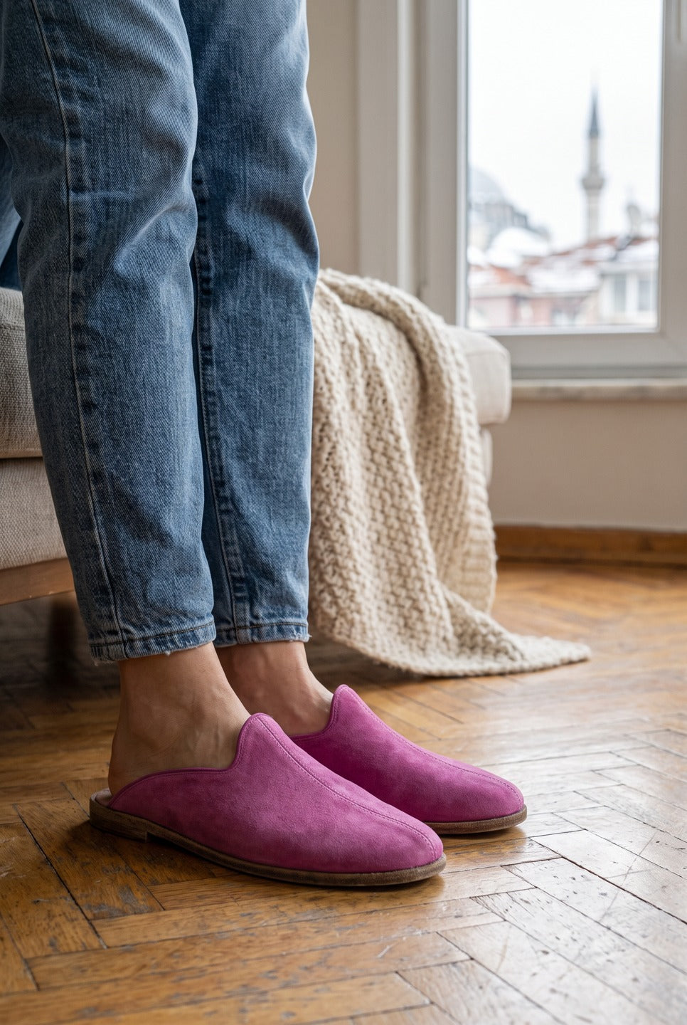 Person in light jeans wearing pink suede barefoot slippers by Bespoky on a herringbone wooden floor next to a knitted throw and a window with a mosque view