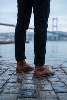 Person in black rolled jeans wearing brown leather barefoot boots by Bespoky on wet cobblestones by the sea with the Bosphorus Bridge in the background