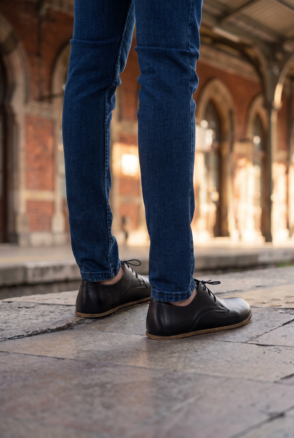 Man in blue jeans wearing black foot-shaped leather oxford shoes by Bespoky on the stone platform of a historic railway station with red brick arches