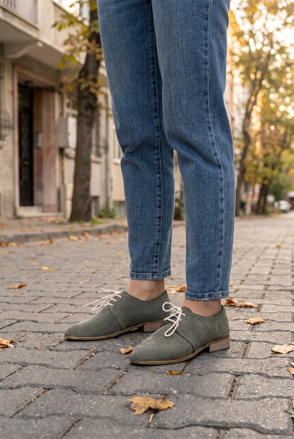 Woman in light blue jeans wearing dark green suede oxford shoes by Bespoky on a cobblestone street with autumn leaves in Istanbul